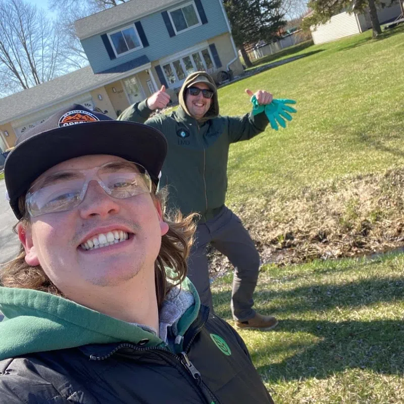 Two men smiling outdoors in a yard with a house in the background on a sunny day.