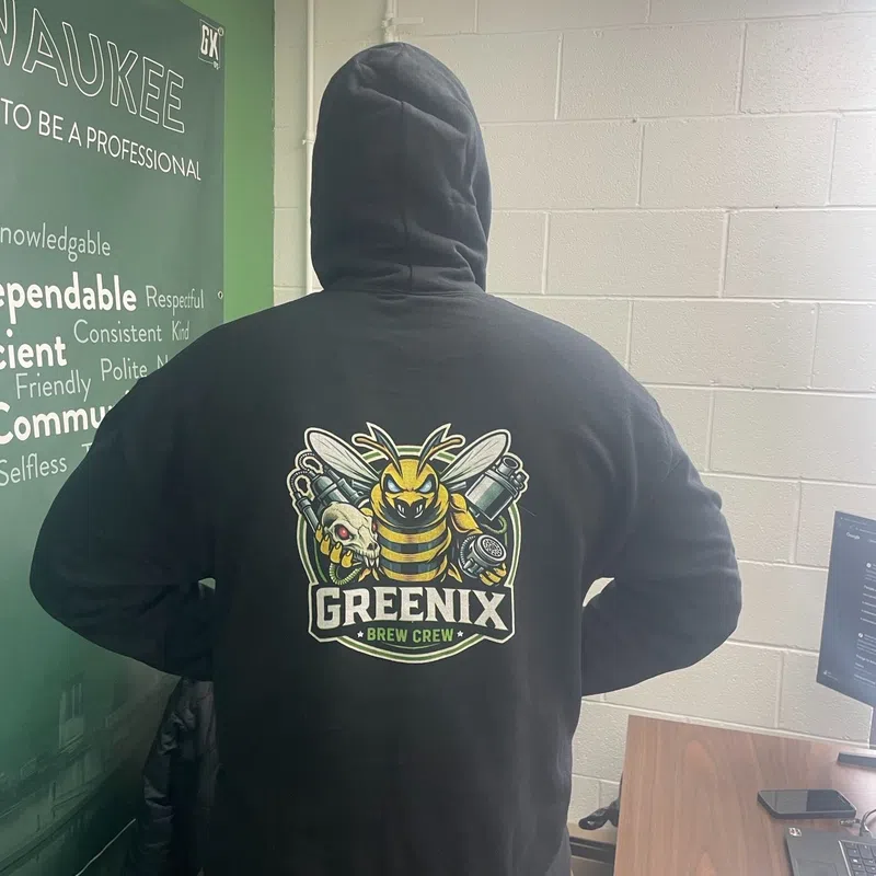 Person wearing black hoodie with Greenix Brew Crew logo featuring a bee holding brewing equipment in an office.