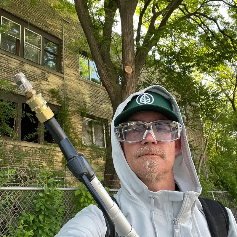 Man in safety glasses and hooded jacket holding a tool outdoors near a brick building and trees.