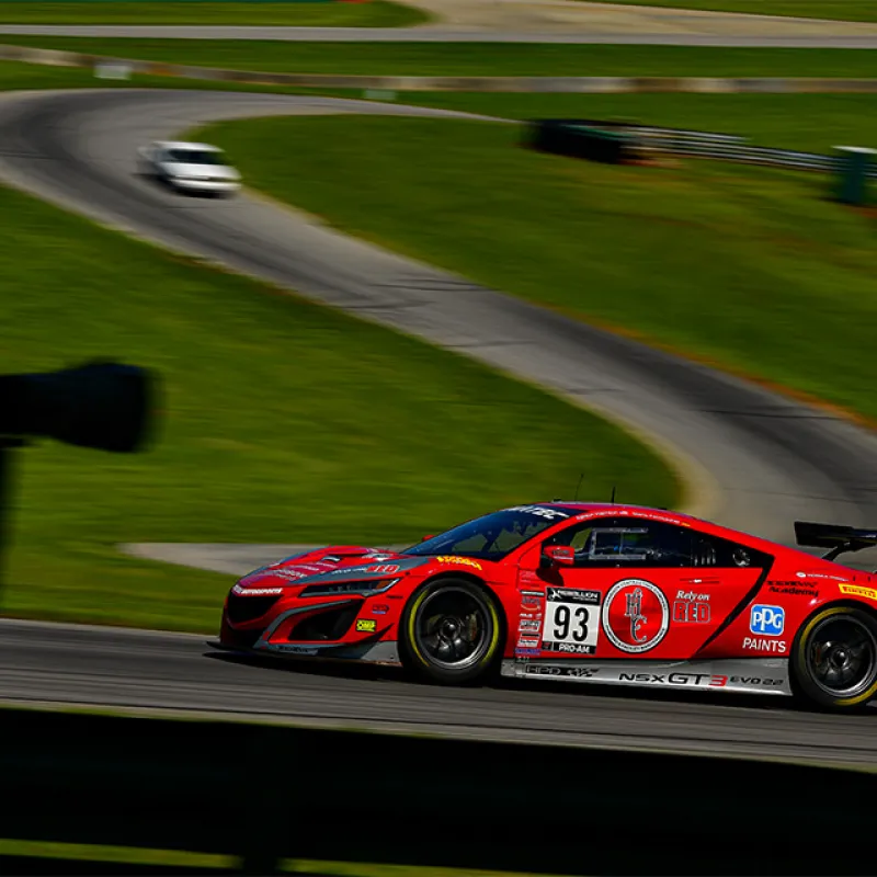 Red race car number 93 speeding on a racetrack with a blurred photographer silhouette in foreground