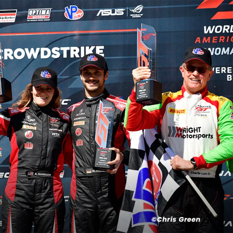 Three race car drivers celebrate on podium holding trophies and checkered flag after GT America race win.