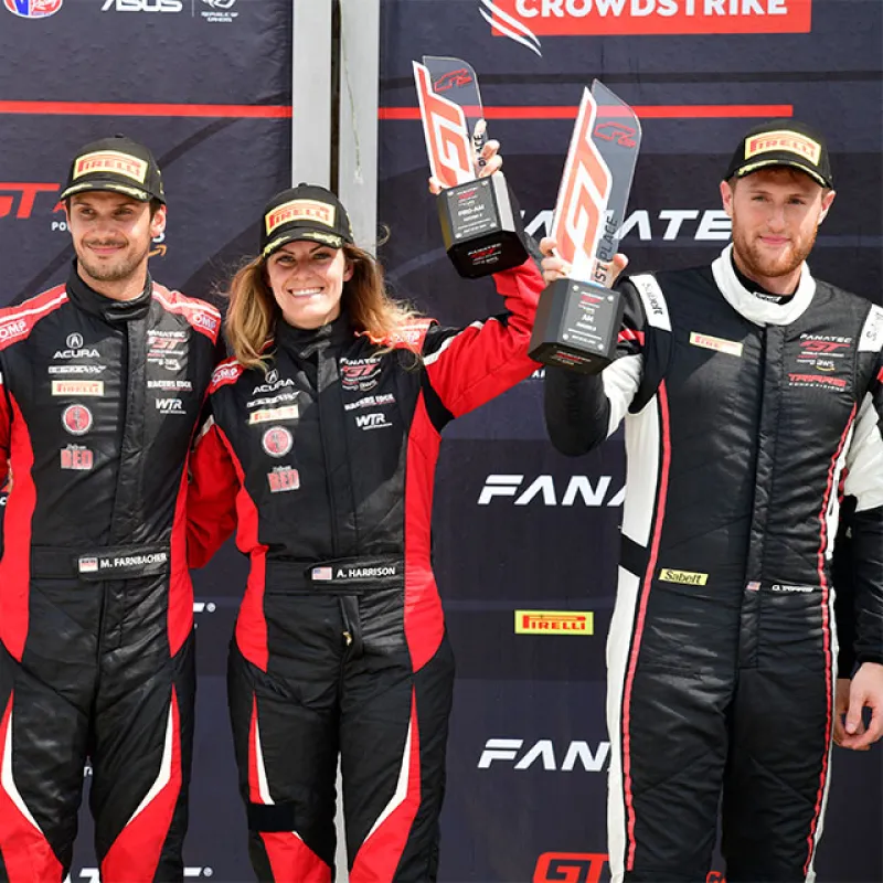 Three race car drivers in black and red racing suits holding trophies on a podium with sponsor banners behind them