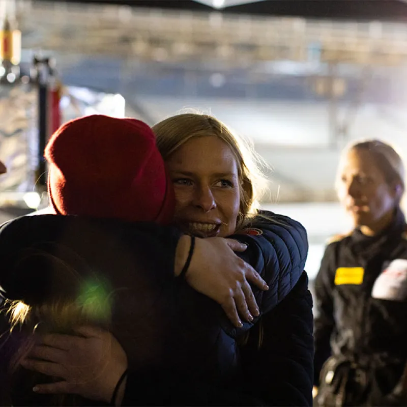 Two women share a warm embrace amid others in racing suits inside a garage with soft lighting.