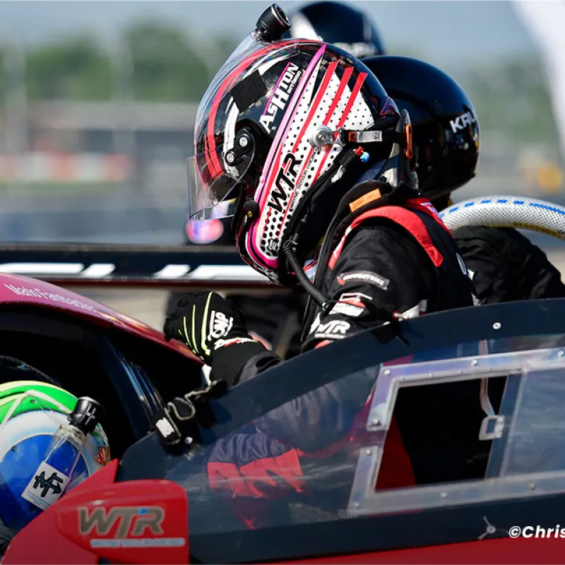 Race car driver wearing helmet and gloves prepares to enter red WTR racing vehicle on track.