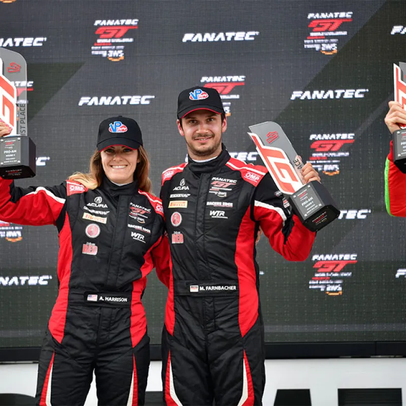 Two race car drivers in red and black suits holding trophies on a Fanatec GT podium with smiles.