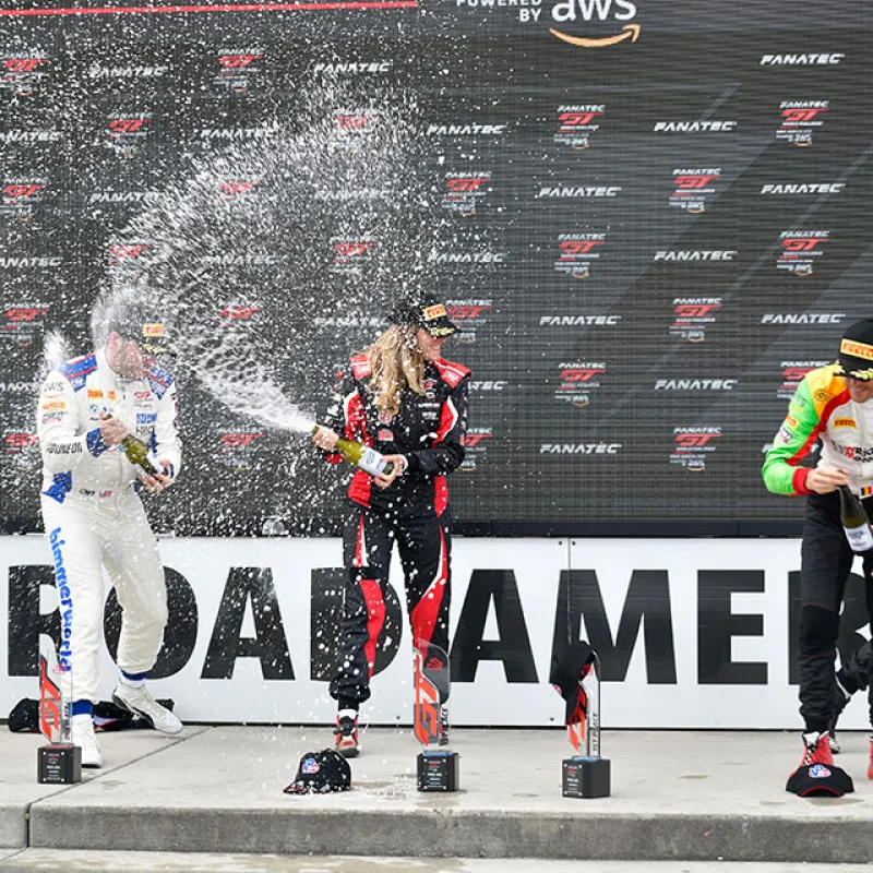 Race car drivers celebrate on podium spraying champagne during motorsport event at Road America circuit.