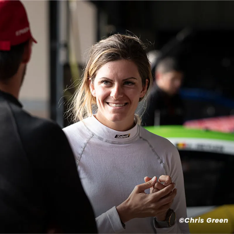 Smiling woman in racing suit talking to people in a garage with race cars in the background