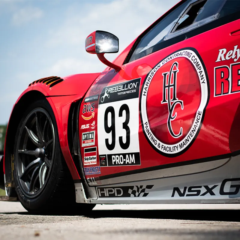 Close-up of a red NSX GT3 race car with number 93 and sponsor logos on a racetrack under blue sky.