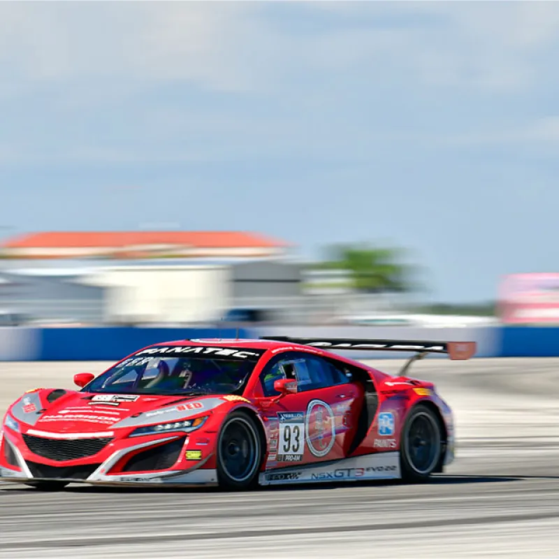 Red Acura NSX GT3 race car speeding on racetrack during a sunny day motorsport event.