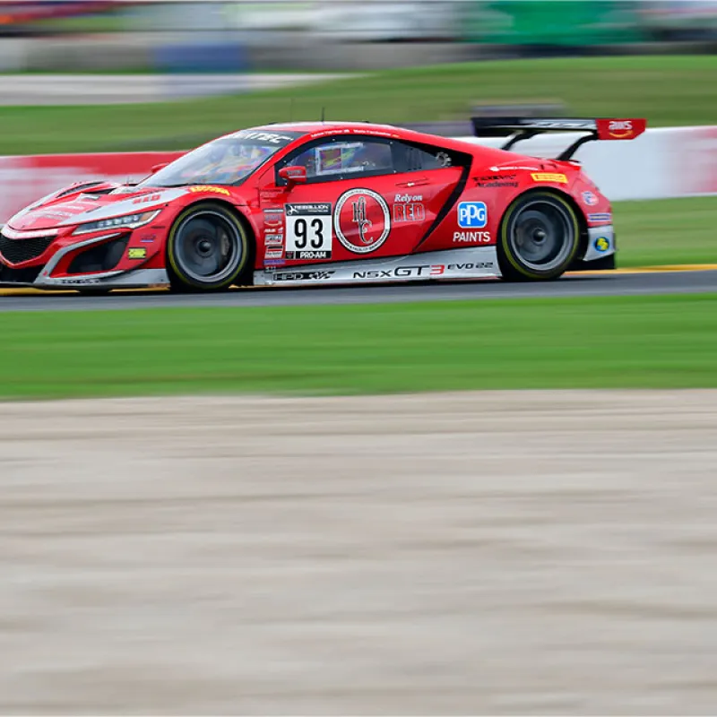 Red Acura NSX GT3 race car number 93 speeding on a green racetrack with blurred background