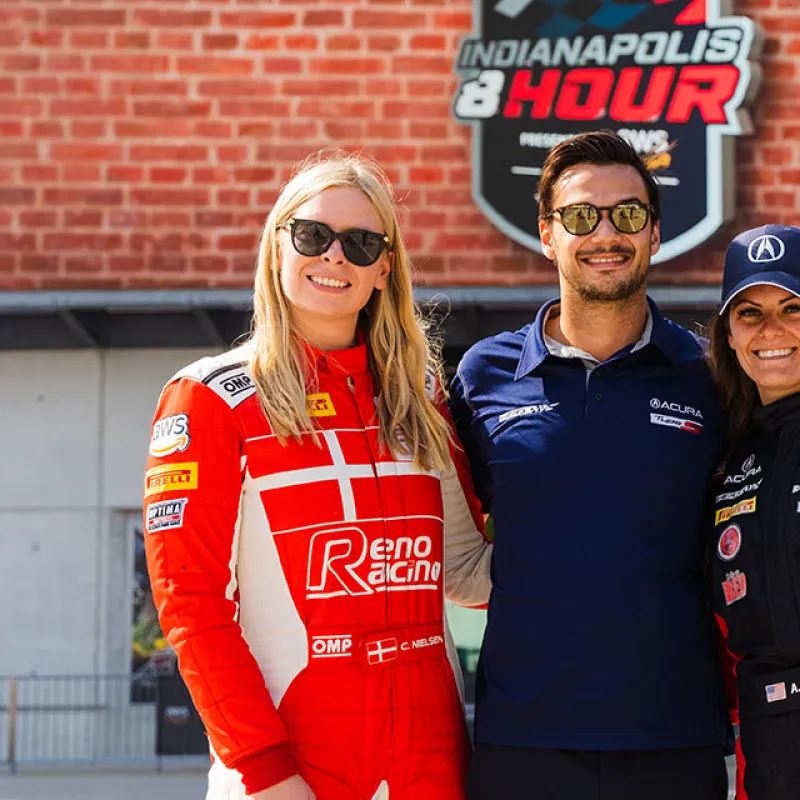 Three smiling race car drivers in racing suits and sunglasses standing in front of Indianapolis 8 Hour event sign.