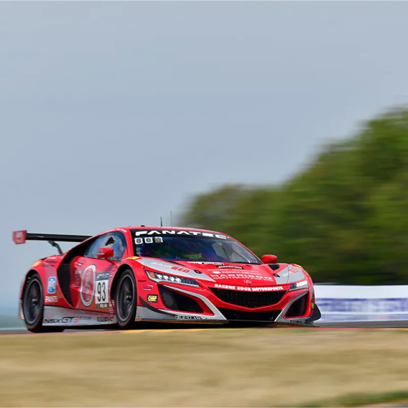Red race car speeding on a racetrack with blurred background and sponsors' logos on the vehicle.