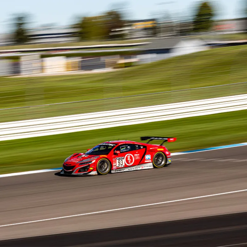 Red race car number 93 speeding on a racetrack with blurred background and green grass.
