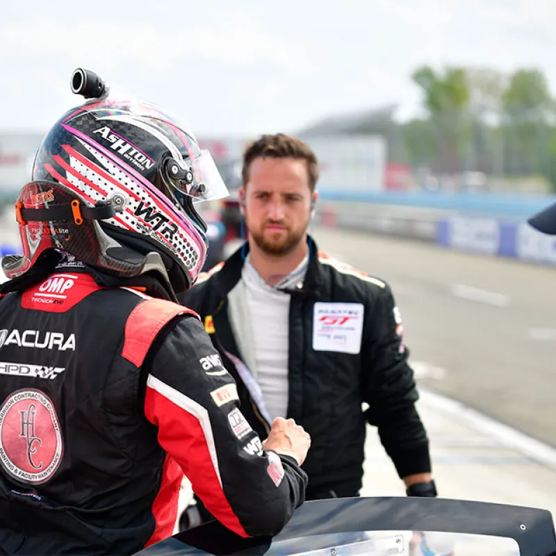 Race car driver in helmet and suit preparing at the pit lane with team member during a motorsport event.