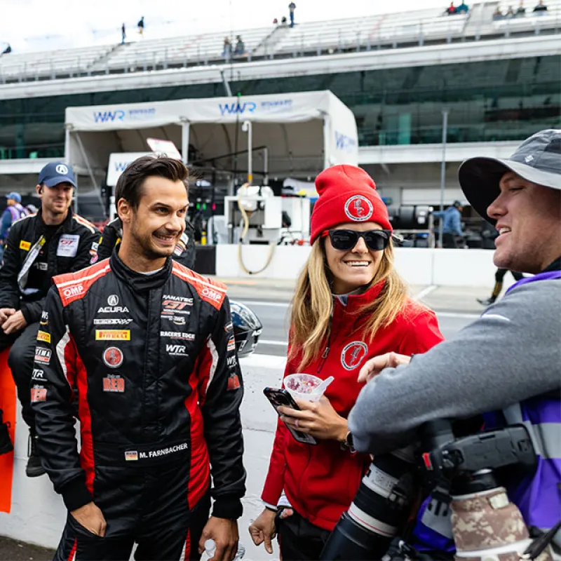 Race car driver in black and red suit smiles while interacting with crew members at a racetrack pit stop.