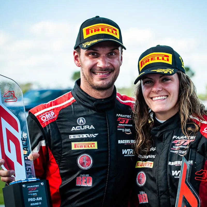 Two race car drivers in black and red suits holding GT racing trophies, smiling with Pirelli caps outdoors.