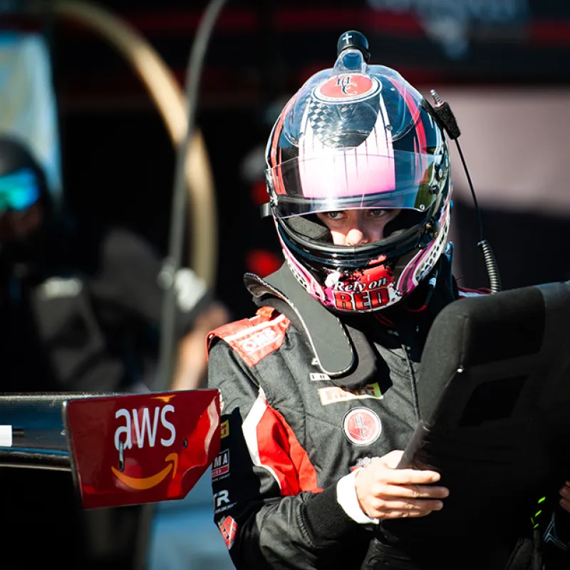 Race car driver in helmet reviewing data on tablet near AWS-branded car wing in pit lane area.