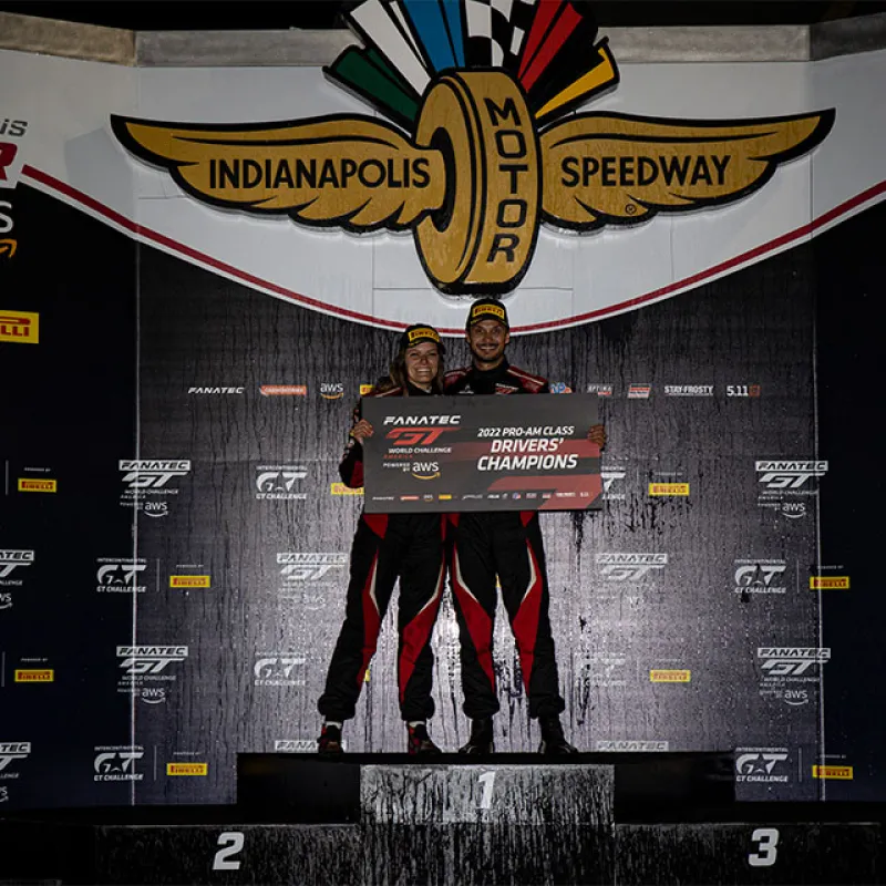 Winning drivers in racing suits celebrate on Indianapolis Motor Speedway podium holding champion banner.