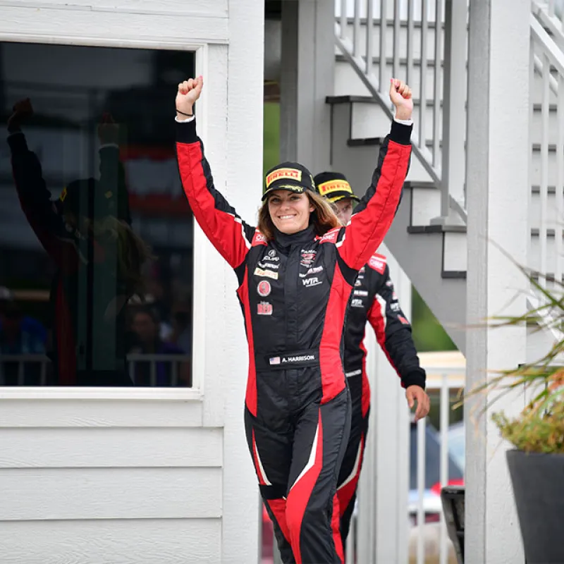Female race car driver in red and black suit raising arms in victory with another driver behind her.