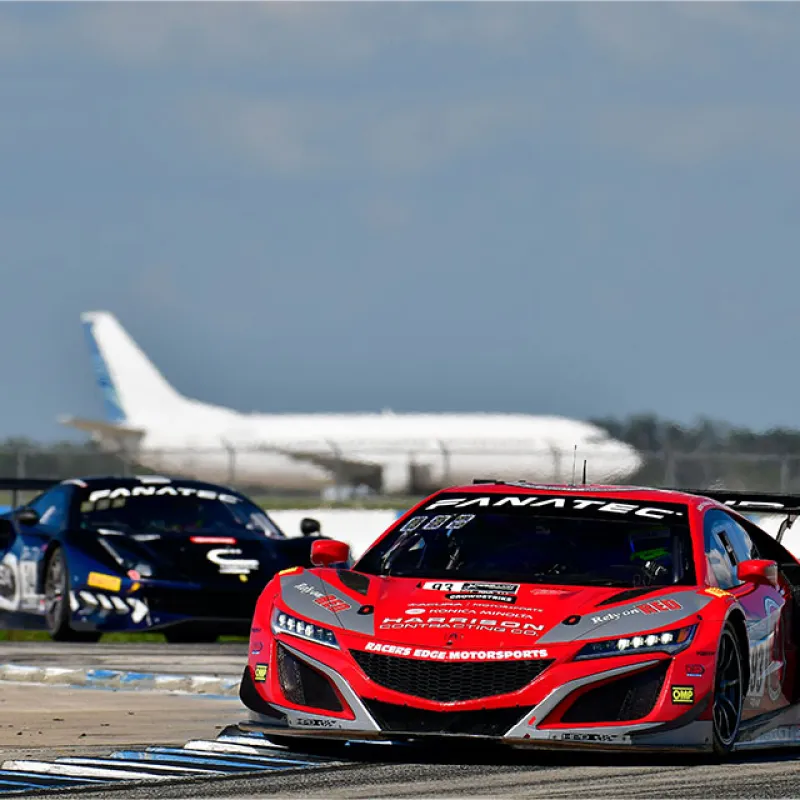 Red and black race cars on a track with a large airplane in the background during a motorsport event.
