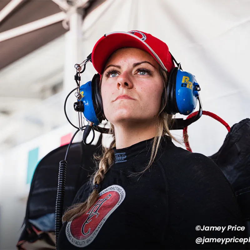 Focused female race engineer wearing red cap and blue headset in a motorsport garage environment.
