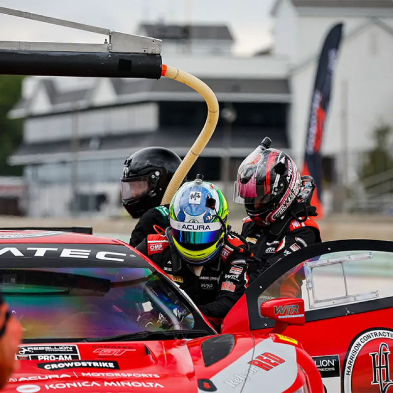 Race car driver in helmet and suit climbing into red Acura VTEC race car during pit stop with crew members nearby.