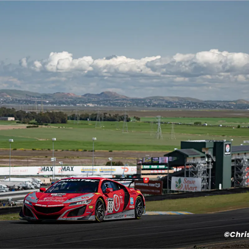 Red race car number 93 speeding on a racetrack with hills and cloudy sky in the background.