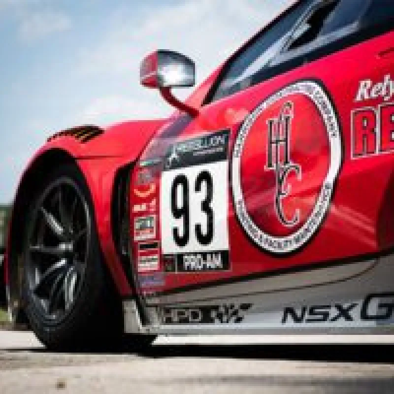 Close-up of red racing car number 93 with sponsor logos and black tire on track under blue sky.