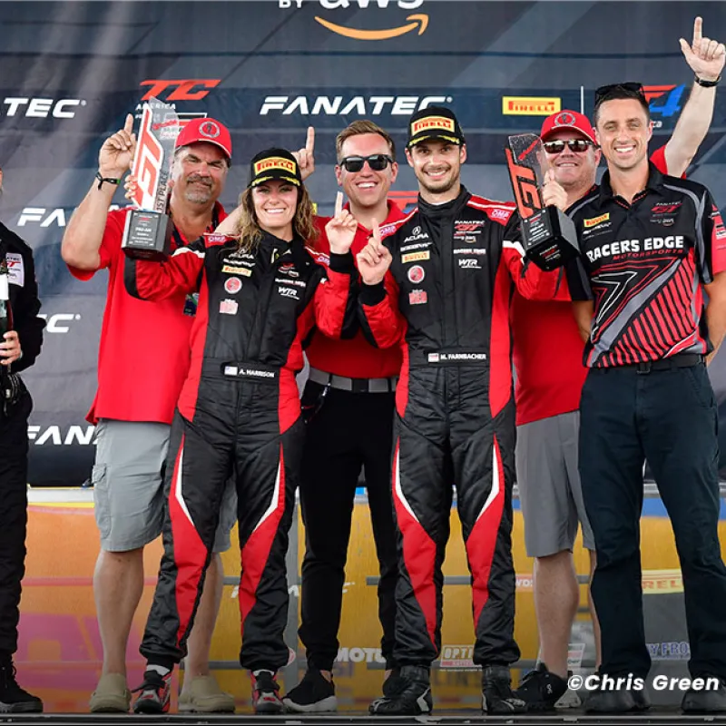 Motorsport team celebrating victory on podium holding trophies and showing number one gestures.