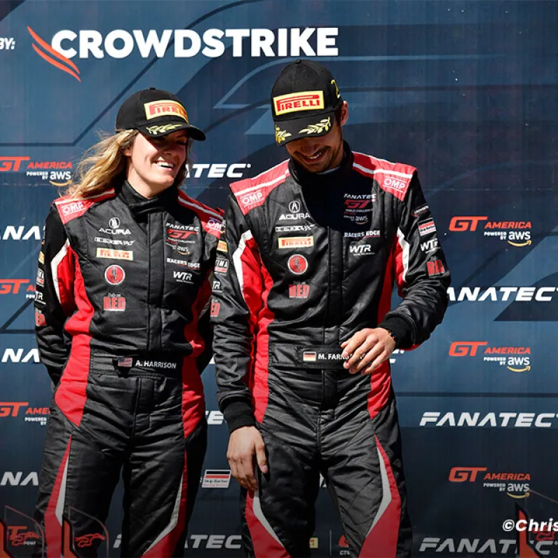 Two race car drivers in black and red racing suits and Pirelli caps standing smiling against a CrowdStrike backdrop.