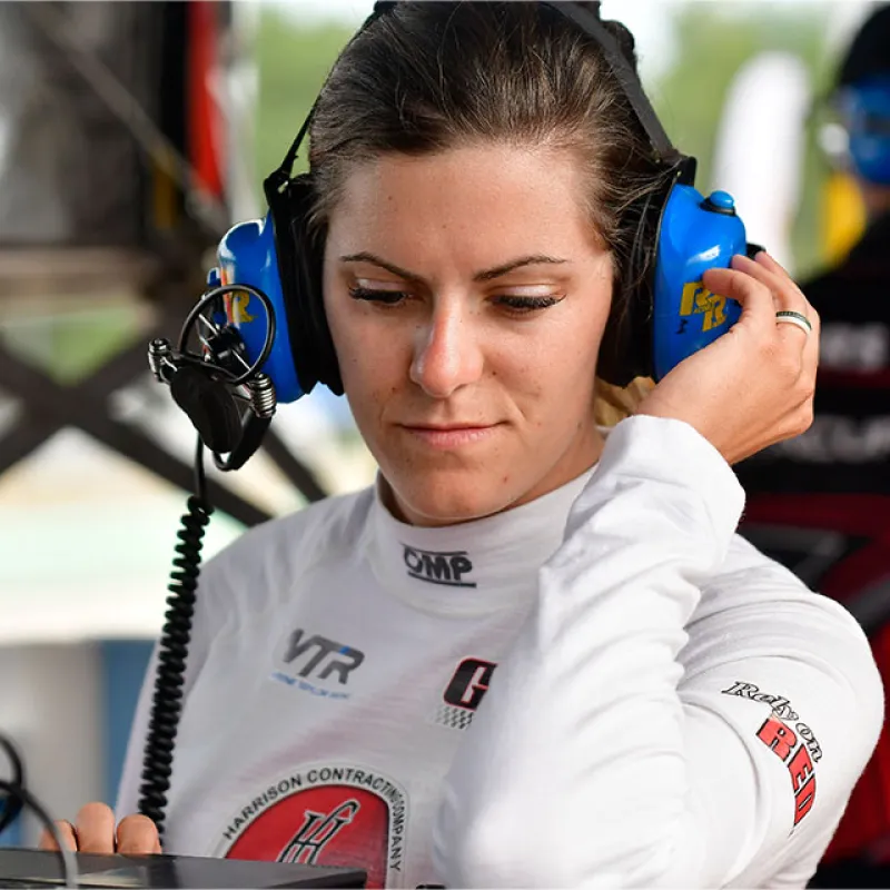 Female race engineer wearing headset and racing suit focused on laptop in motorsport pit area