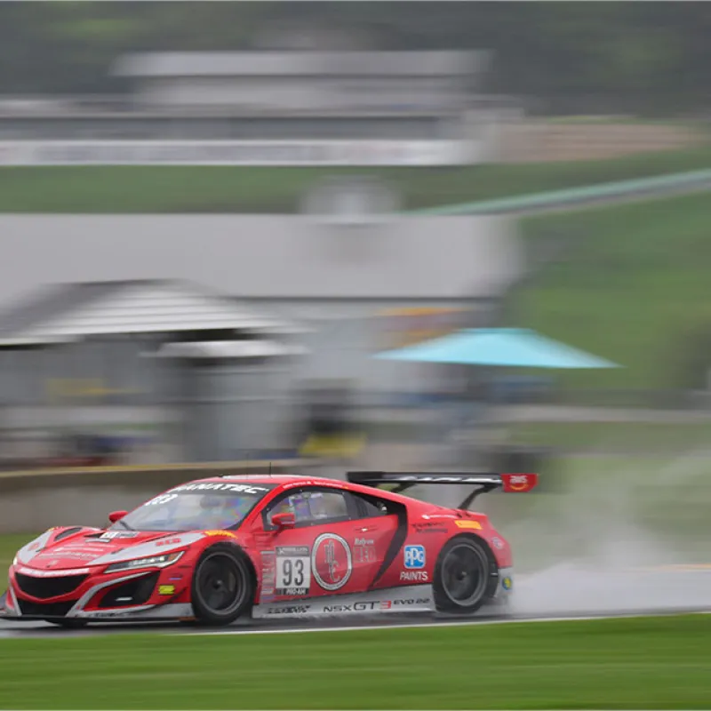 Red Acura NSX GT3 race car number 93 speeding on wet race track with spray and blurred background