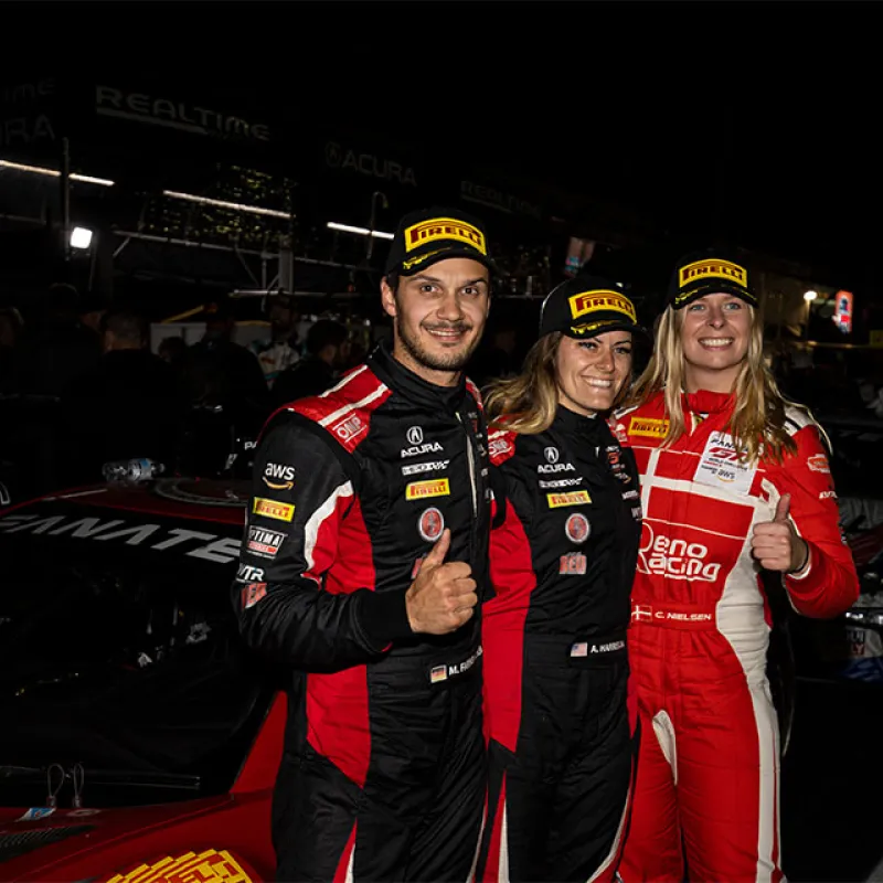 Three race car drivers in racing suits and Pirelli hats standing by a race car at night, smiling and giving thumbs up.