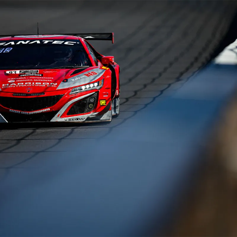 Red race car with sponsors speeding on a racetrack near the wall under sunlight.