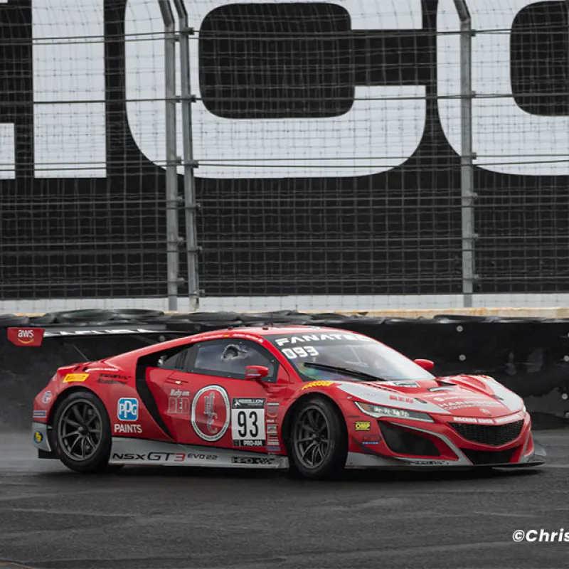 Red Acura NSX GT3 race car number 93 drifting on wet track with sponsor decals in front of racetrack barrier.