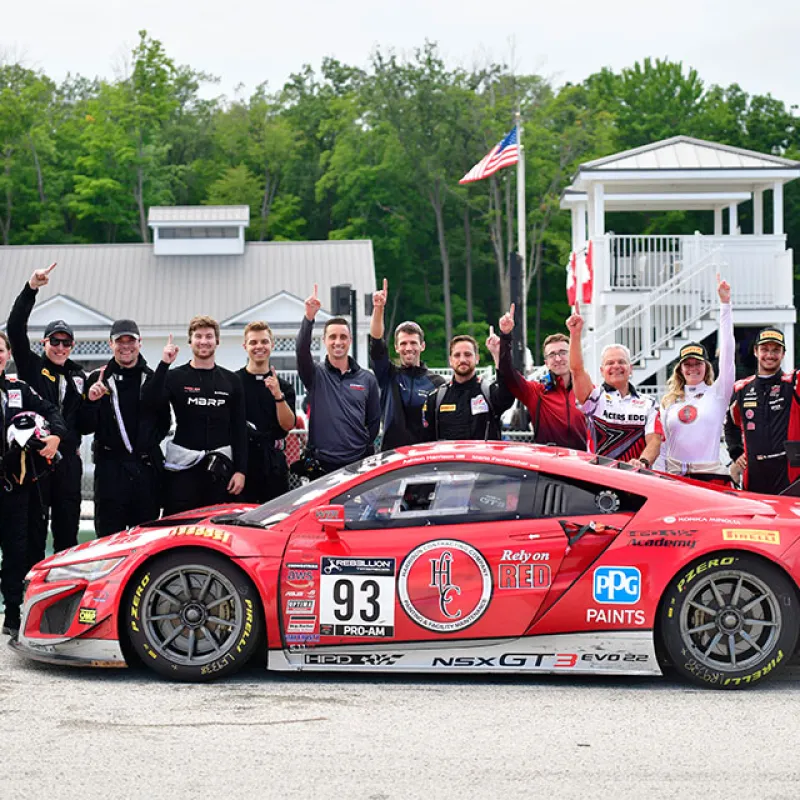 Race team celebrates next to red Acura NSX GT3 EVO 22 race car at track with flags and greenery behind