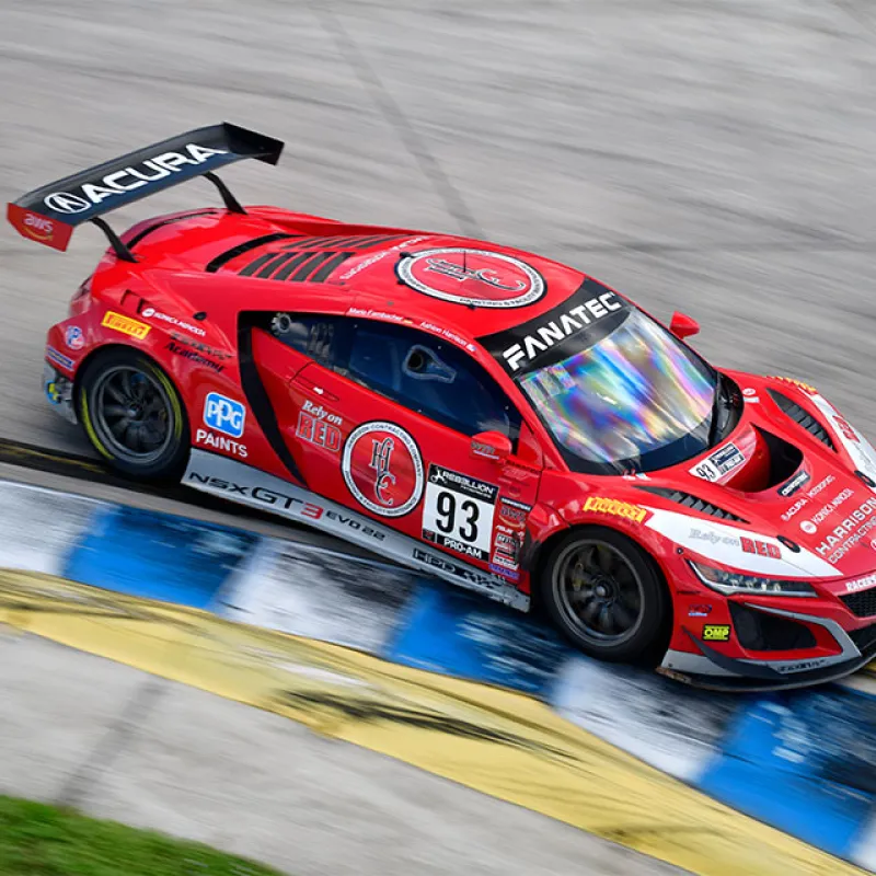 Red Acura racing car number 93 speeding on a racetrack with sponsor decals and aerodynamic wing.