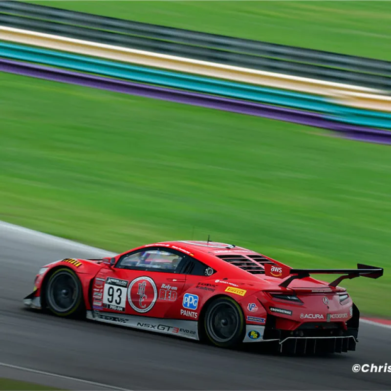 Red Acura NSX GT3 race car number 93 speeding on a racetrack with blurred green and purple background.