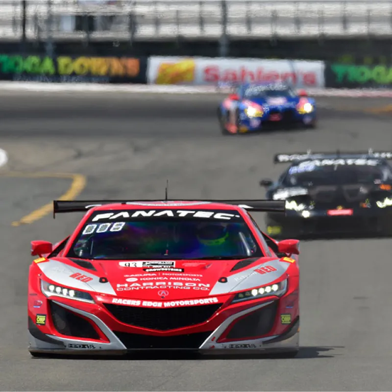 Red and black race cars competing on a track during a high-speed motorsport event with sponsorship banners in background