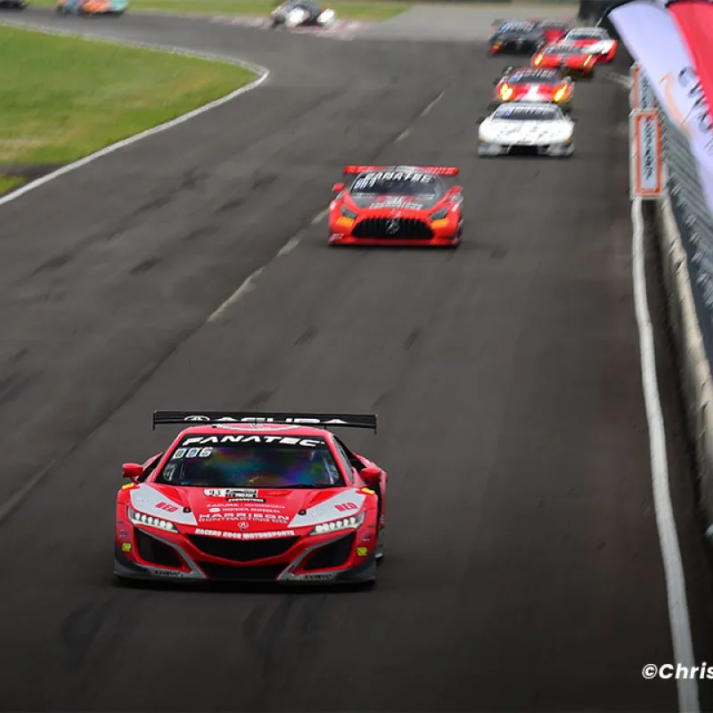Red race car leading a pack on a racetrack with flags and safety barriers alongside the track.
