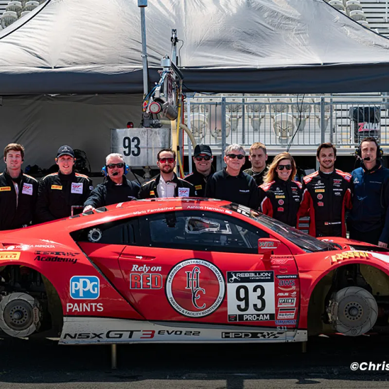 Red NSX GT3 EVO 22 race car number 93 with crew posing in front of pit garage under canopy at race track.