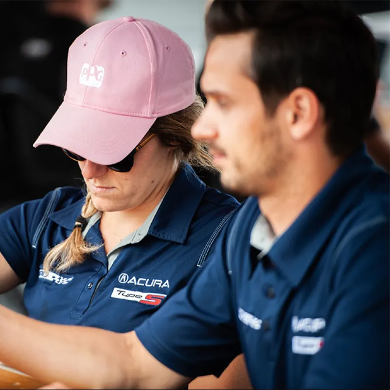 Woman in pink cap and Acura shirt writes at a desk beside a man in similar uniform during an event.
