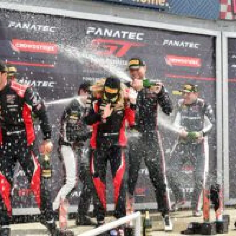Race car drivers celebrate on podium spraying champagne during race victory ceremony with branded backdrop