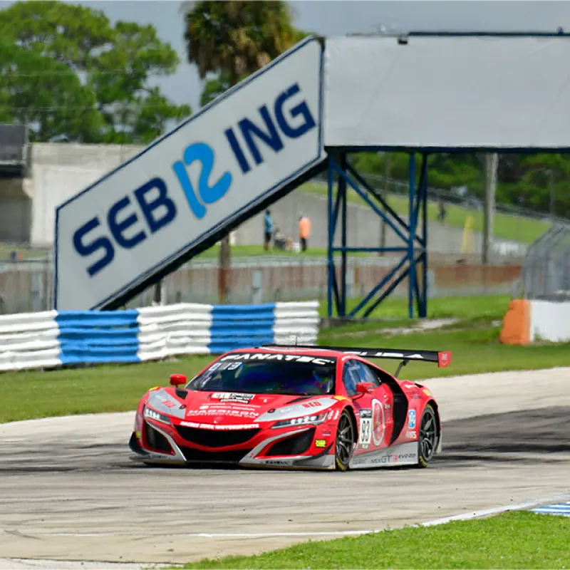 Red race car speeding on Sebring raceway track with blue and white curbs under sunny sky.