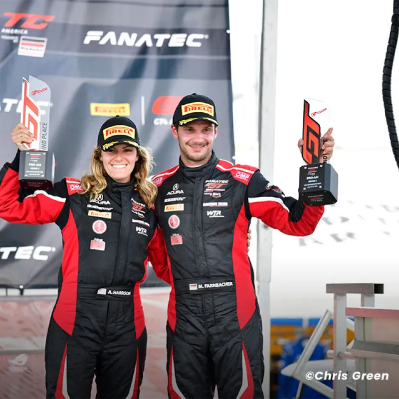 Two race car drivers in black and red suits hold GT trophies and smile on the Fanatec podium