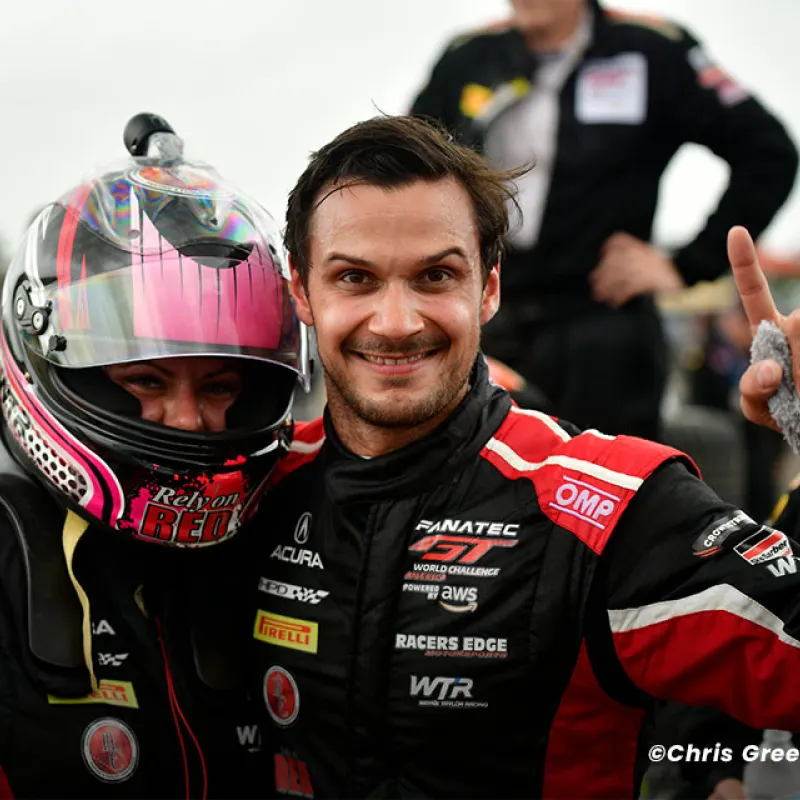 Two race car drivers in black and red racing suits celebrating with raised index fingers, one wearing a pink helmet.