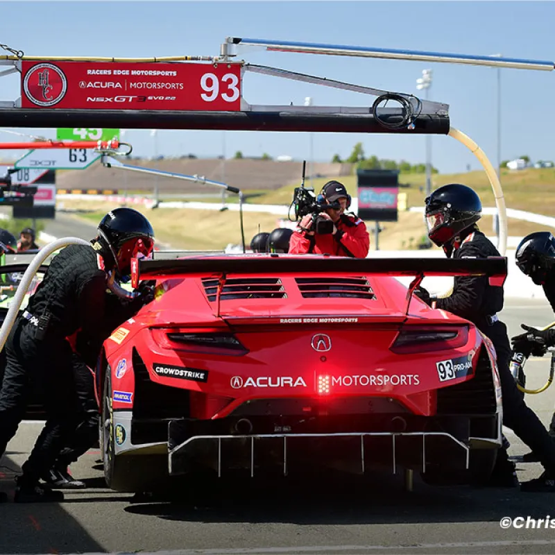 RACER'S EDGE Acura red racecar in a pit stop with crew members refueling and changing tires on the track.