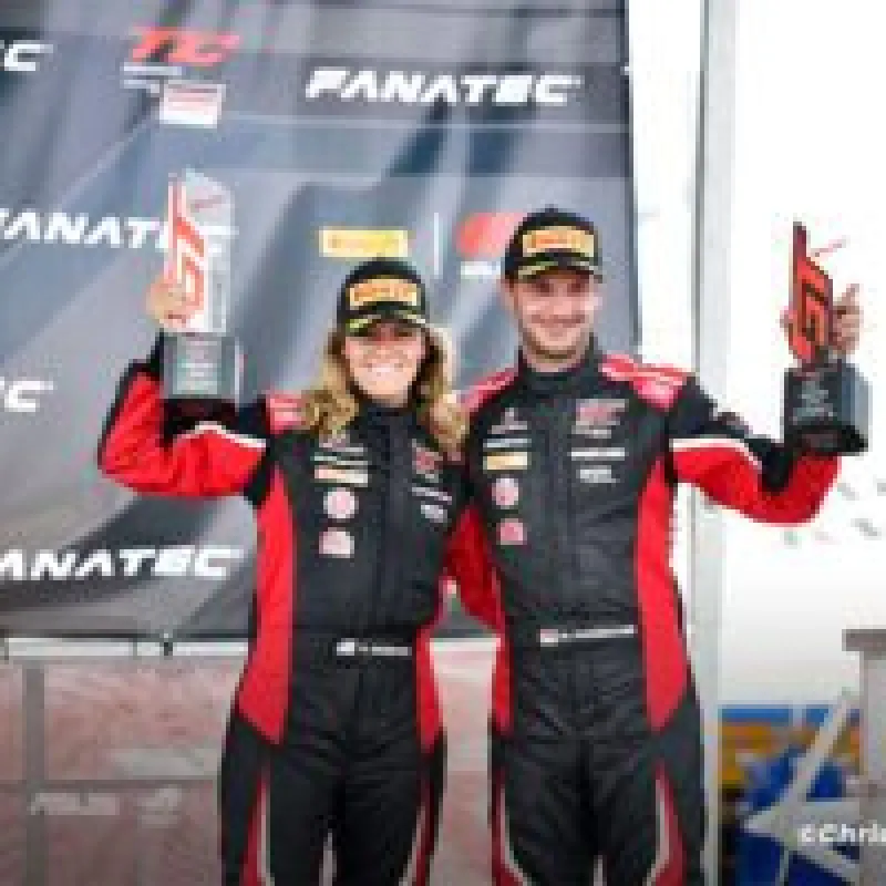 Two race car drivers in black and red suits holding trophies on a motorsport podium with Fanatec branding.