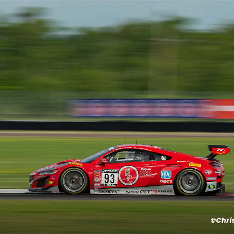 Red GT3 race car number 93 speeds on racetrack with blurred green background during daytime race.