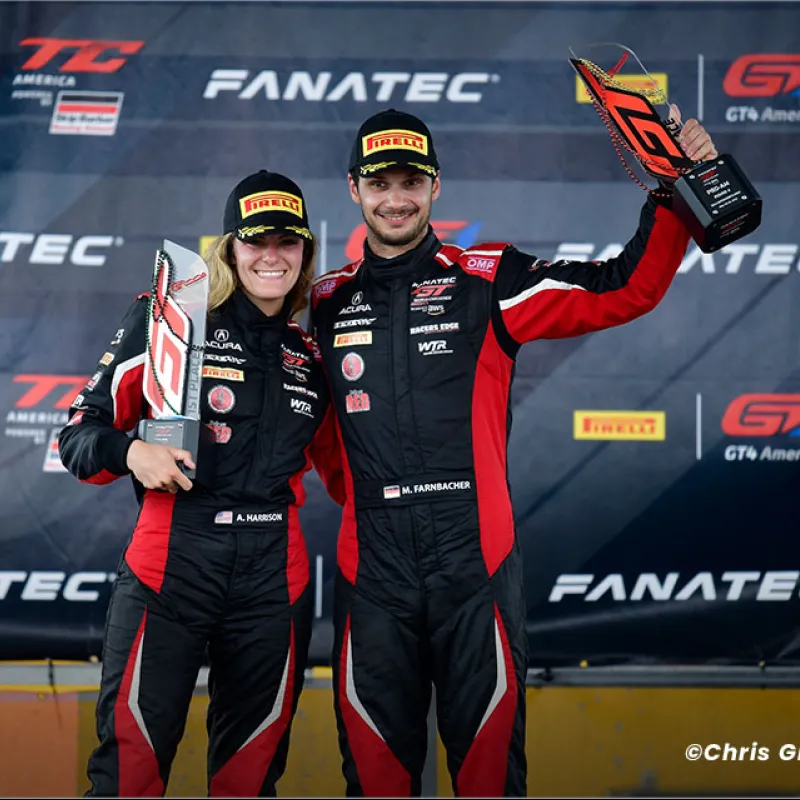 Two race car drivers in black and red suits holding trophies on a podium at a Fanatec GT4 America event